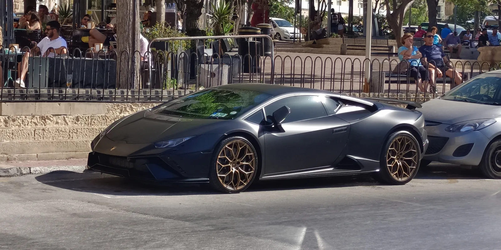 A dark grey Lamborghini Huracán parked on a palm-lined seafront street in Malta, next to an ordinary hatchback