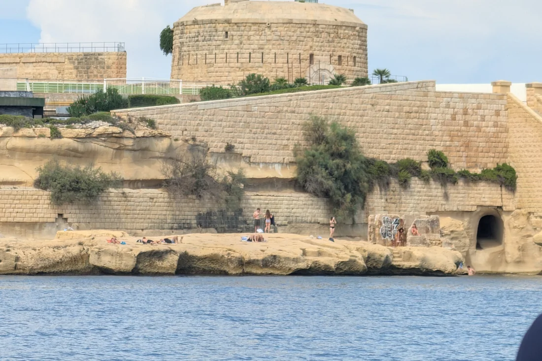 A coastal fort seen from the water near the Comino crossing, with people on the rocks below