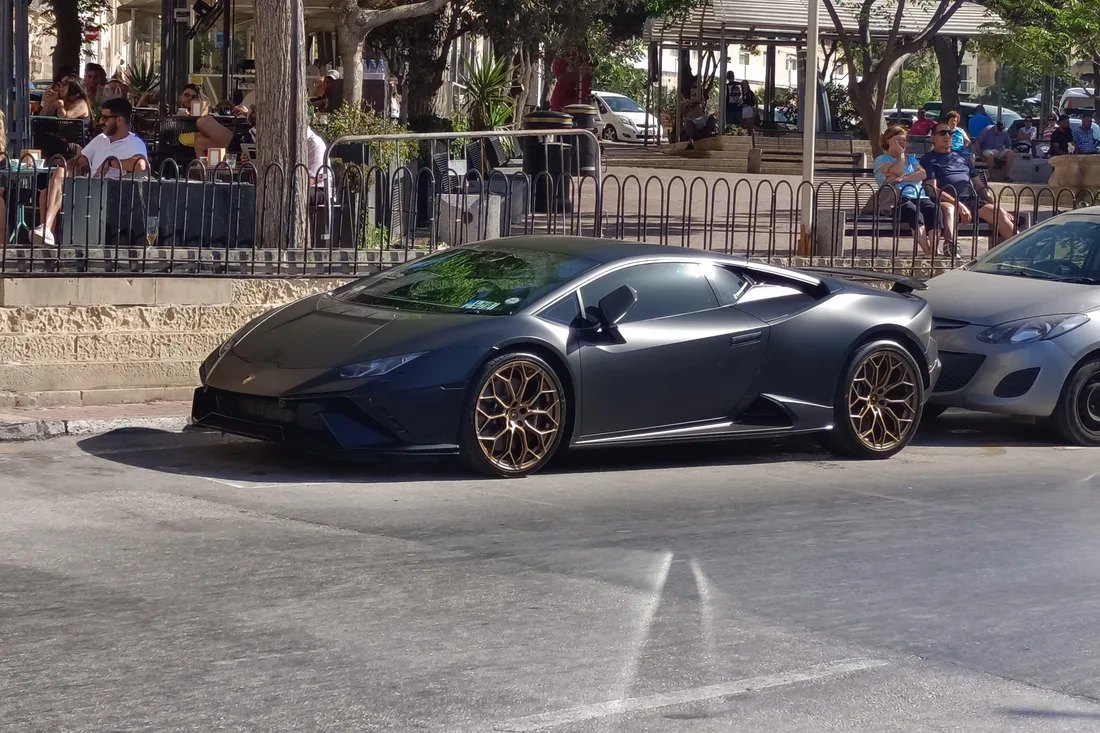A dark grey Lamborghini Huracán parked on a tree-lined seafront street in Malta, next to an ordinary hatchback