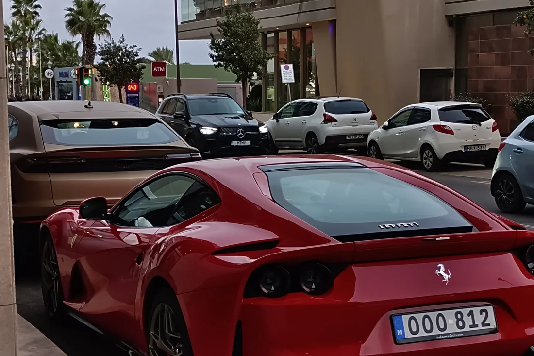 A red Ferrari 812 stuck in evening traffic on a Sliema street, boxed in by ordinary hatchbacks